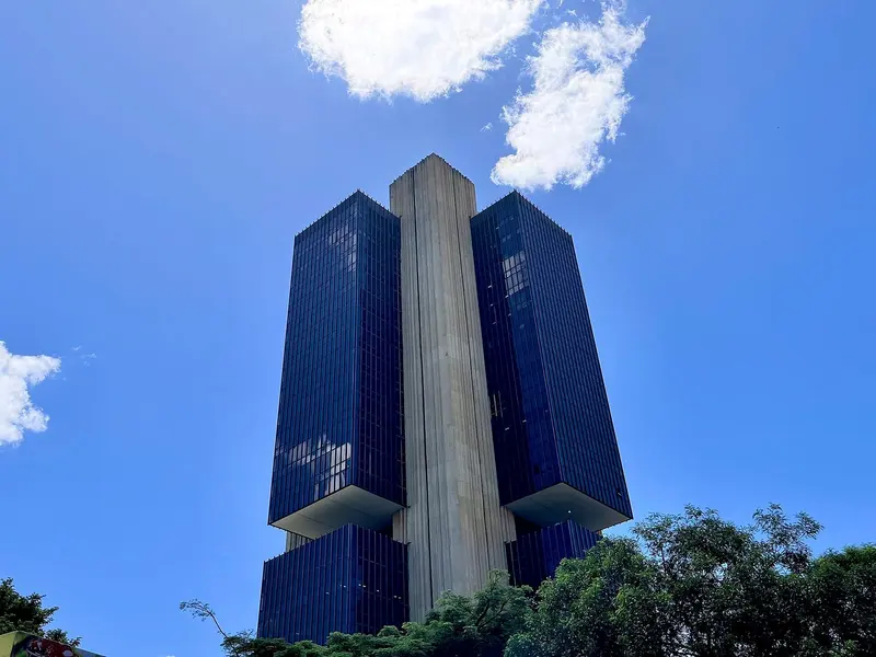 Sede do Banco Central em Brasília: dois blocos de vidro escuro ao lado de uma torre central de concreto, sob céu azul